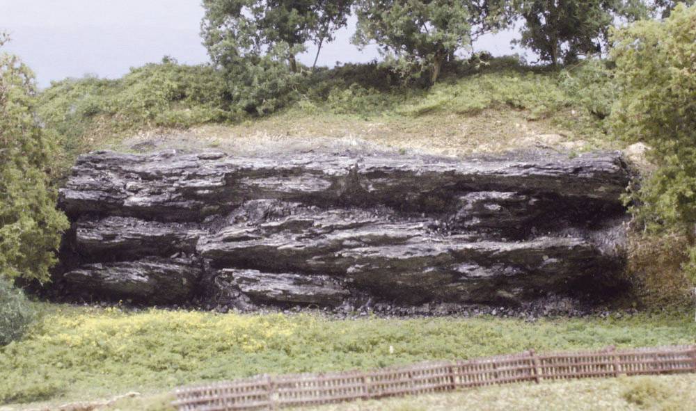 Felsformation in einer grünen Landschaft mit Bäumen und einem Holzzaun im Vordergrund.
