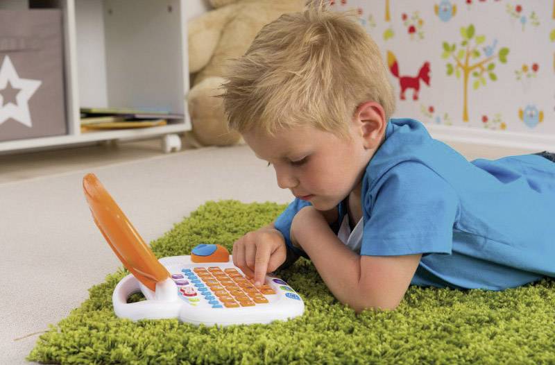 Ein Junge liegt auf einem Teppich und spielt konzentriert mit einem Lernspielzeug-Laptop. Im Hintergrund ist Kinderzimmerdekoration zu sehen.