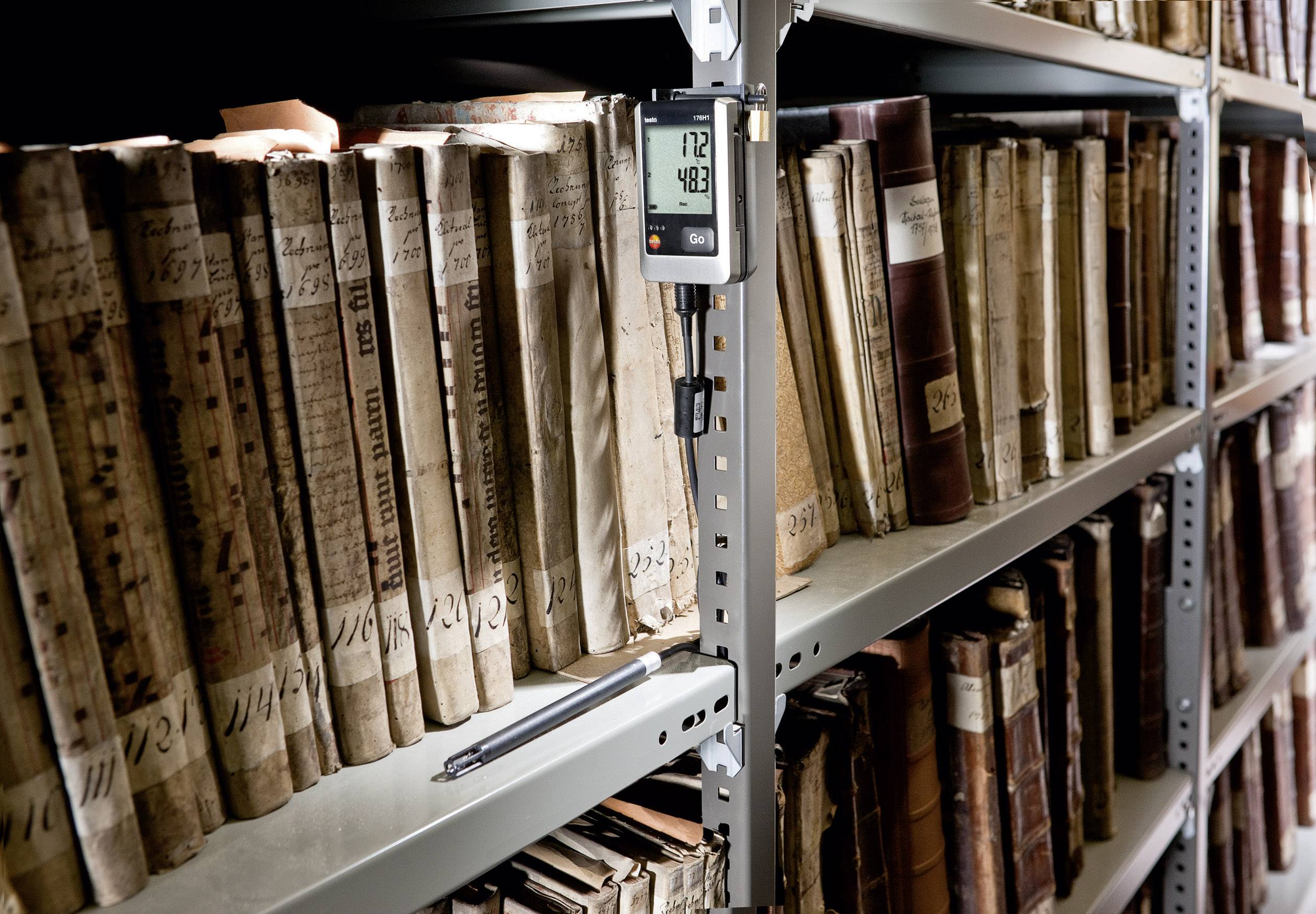 Regale mit alten Büchern in einer Bibliothek, ein Thermohygrometer zeigt '12°C' und '48%' an, Stift liegt auf dem Regal.