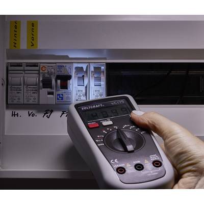 Eine Hand hält ein Digitalmultimeter vor einem offenen Sicherungskasten, um die Spannung zu messen. Die Anzeige zeigt '234.9' an.