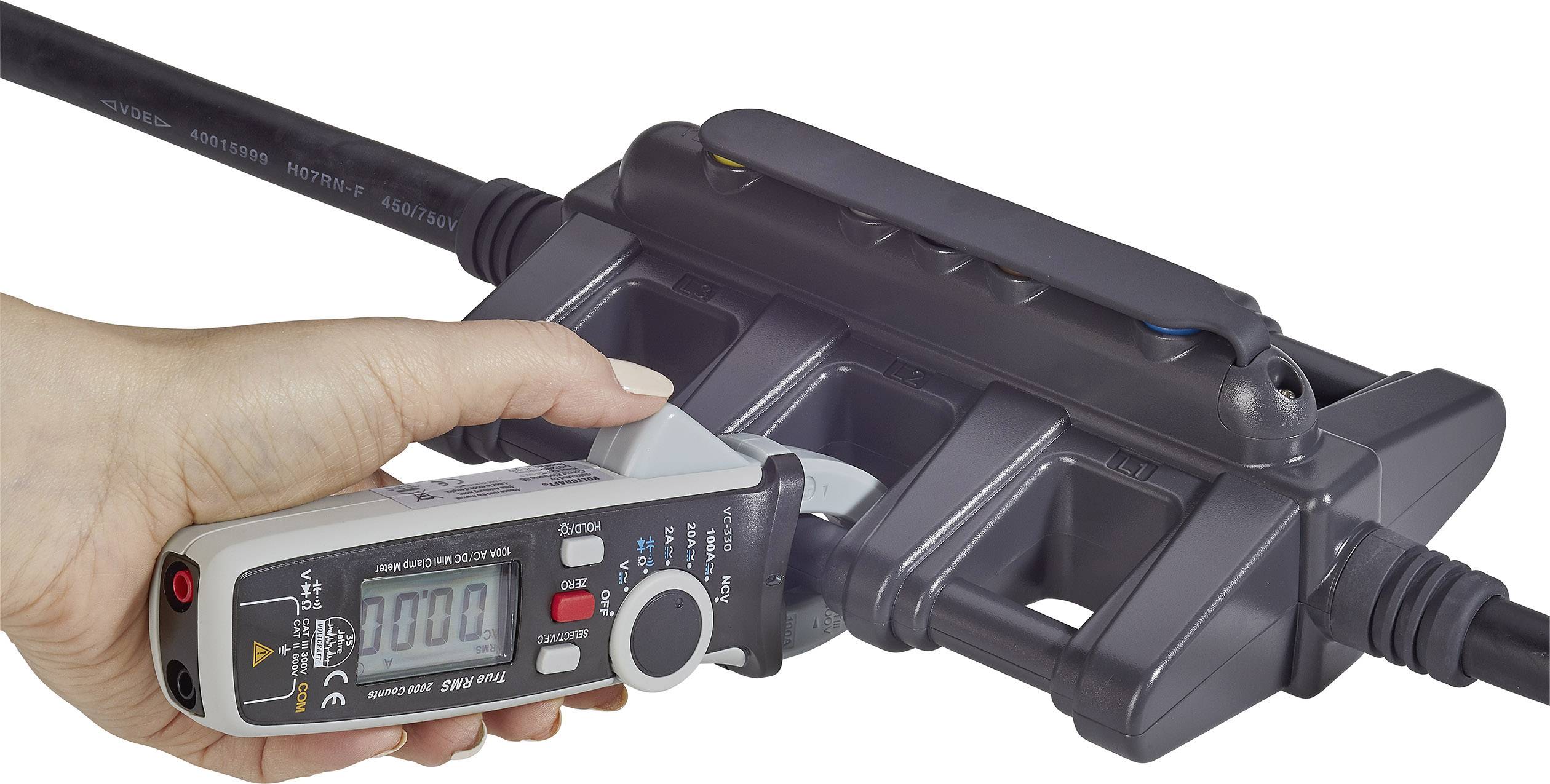 Eine Hand hält ein digitales Multimeter, das den Strom einer Steckdosenleiste misst. Die Anzeige zeigt '100.0' an.