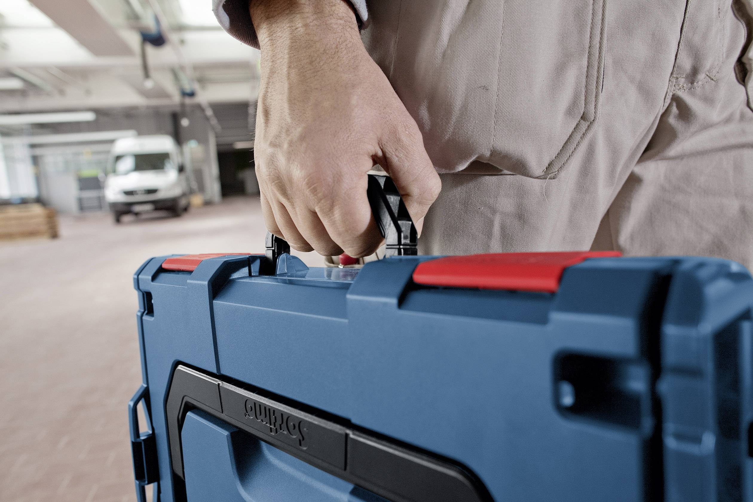 Eine Person in Arbeitskleidung trägt einen blauen Werkzeugkoffer in einem industriellen Innenraum. Im Hintergrund steht ein Lieferwagen.