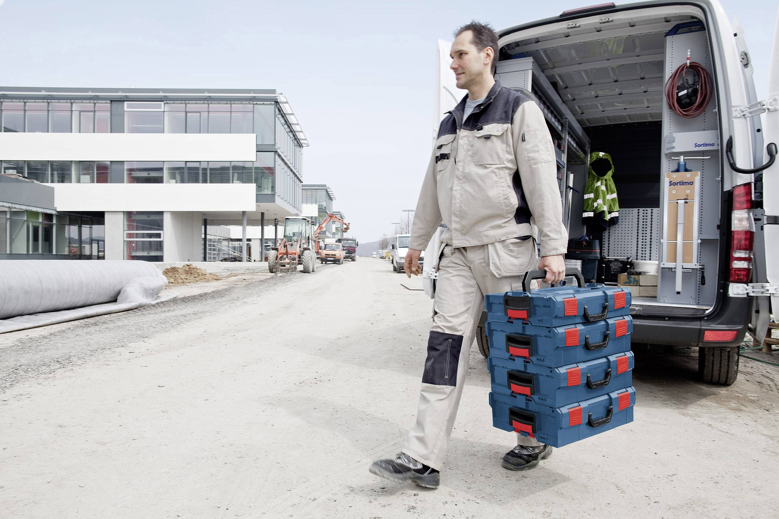 Ein Bauarbeiter trägt stapelbare Werkzeugkoffer und geht an einem Baugelände vorbei, während neben ihm ein Lieferwagen mit offenem Heck steht.