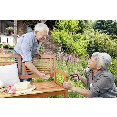 Ein älteres Paar repariert eine Holzbank im Garten. Der Mann hält ein Teil, während die Frau mit einem Akkuschrauber arbeitet. Im Hintergrund blühen Blumen.