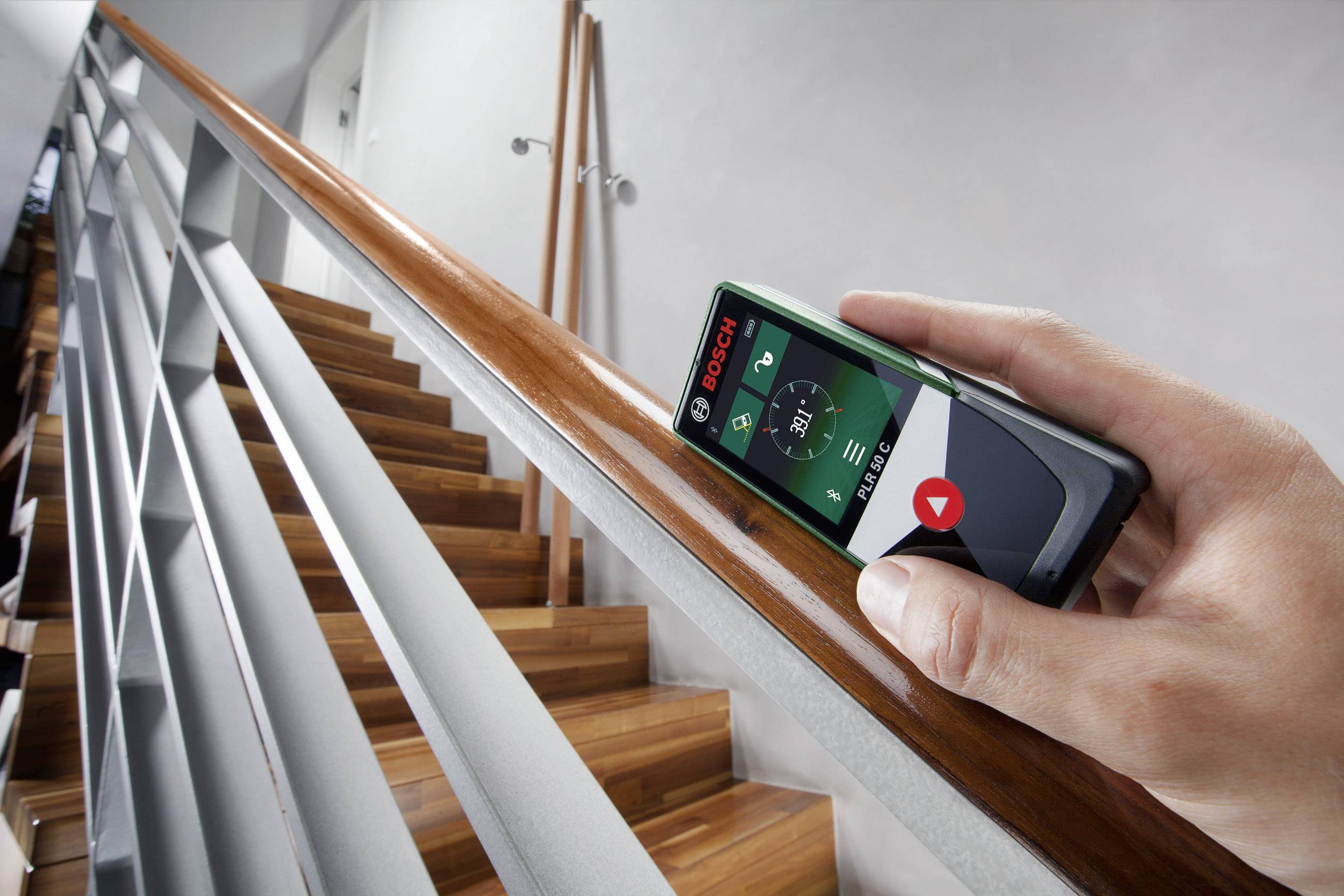 Eine Person misst die Länge eines Holzgeländers mit einem digitalen Laser-Entfernungsmesser auf einer Treppe.