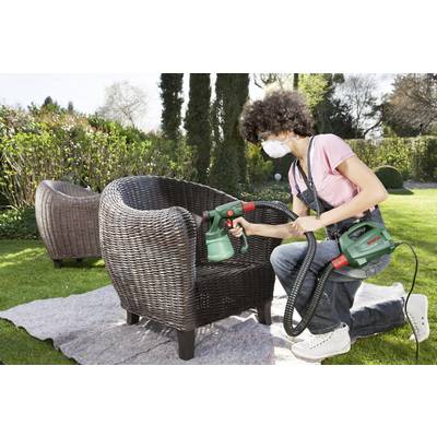 Eine Person sprüht Farbe auf einen Gartenstuhl, trägt dabei Schutzbrille und Maske. Im Hintergrund befindet sich ein Garten.