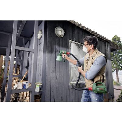 Ein Mann in Schutzbrille und Maske lackiert eine Holzwand mit einem Farbsprüher. Im Hintergrund ist ein Holzhaus zu sehen.