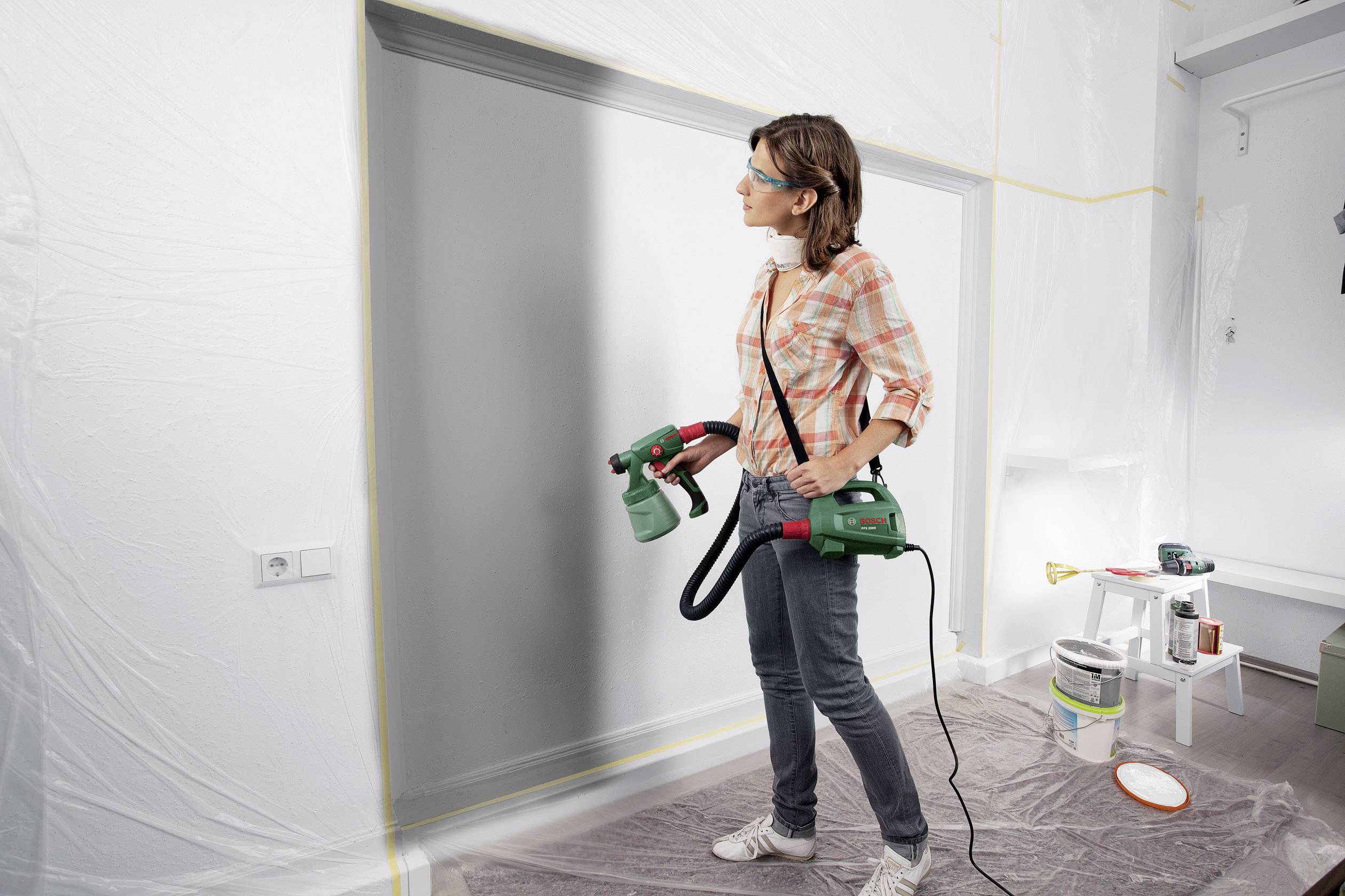 Eine Frau verwendet eine elektrische Farbspritzpistole, um eine Wand grau zu streichen. Die Umgebung ist mit Abdeckfolie geschützt.