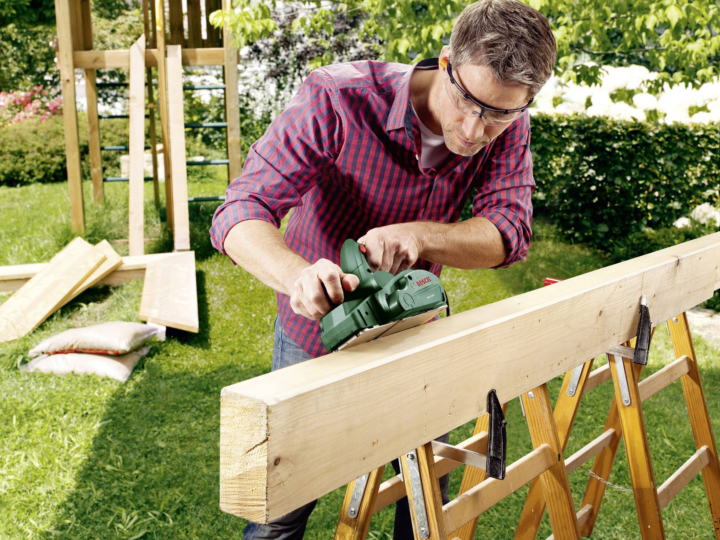 Ein Mann mit Schutzbrille schleift ein Holzbrett auf einer Säge mit einer elektrischen Schleifmaschine in einem Garten.