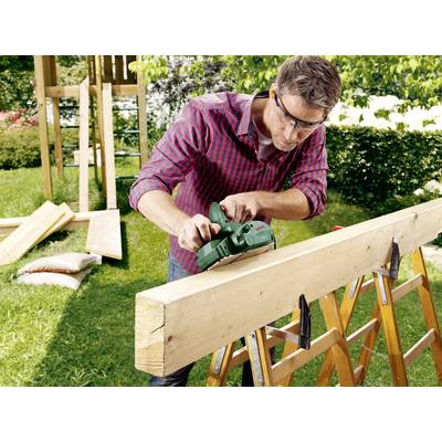 Ein Mann mit Schutzbrille schleift ein Holzbrett auf einer Säge mit einer elektrischen Schleifmaschine in einem Garten.