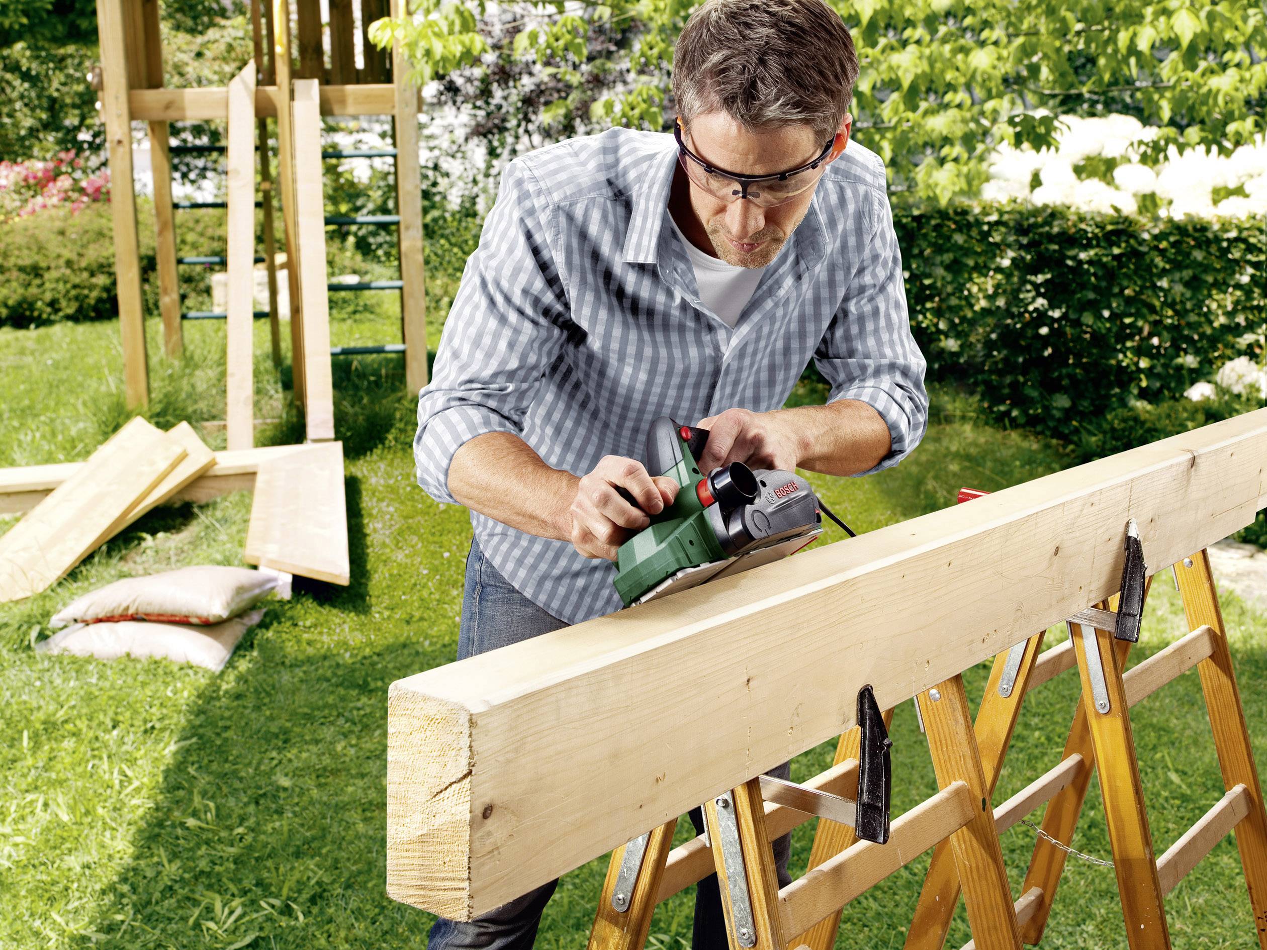 Ein Mann trägt Schutzbrille und benutzt ein Elektrowerkzeug, um ein großes Holzbrett im Garten zu bearbeiten.
