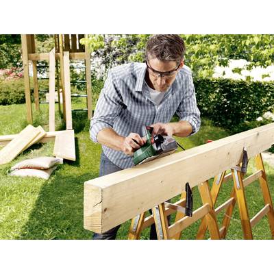 Ein Mann trägt Schutzbrille und benutzt ein Elektrowerkzeug, um ein großes Holzbrett im Garten zu bearbeiten.