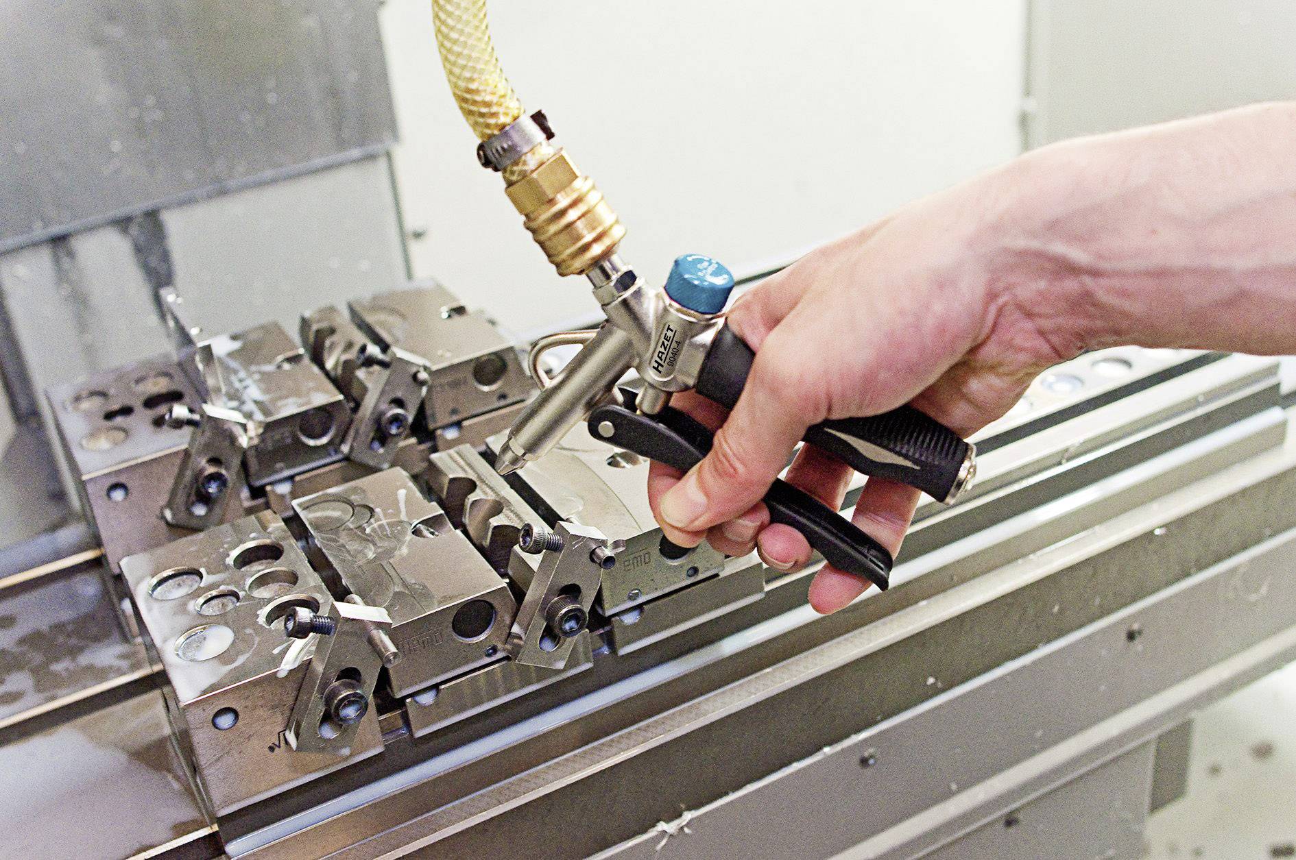 Eine Hand hält eine Druckluftpistole und reinigt metallene Maschinenteile auf einem Tisch in einer Werkstatt.