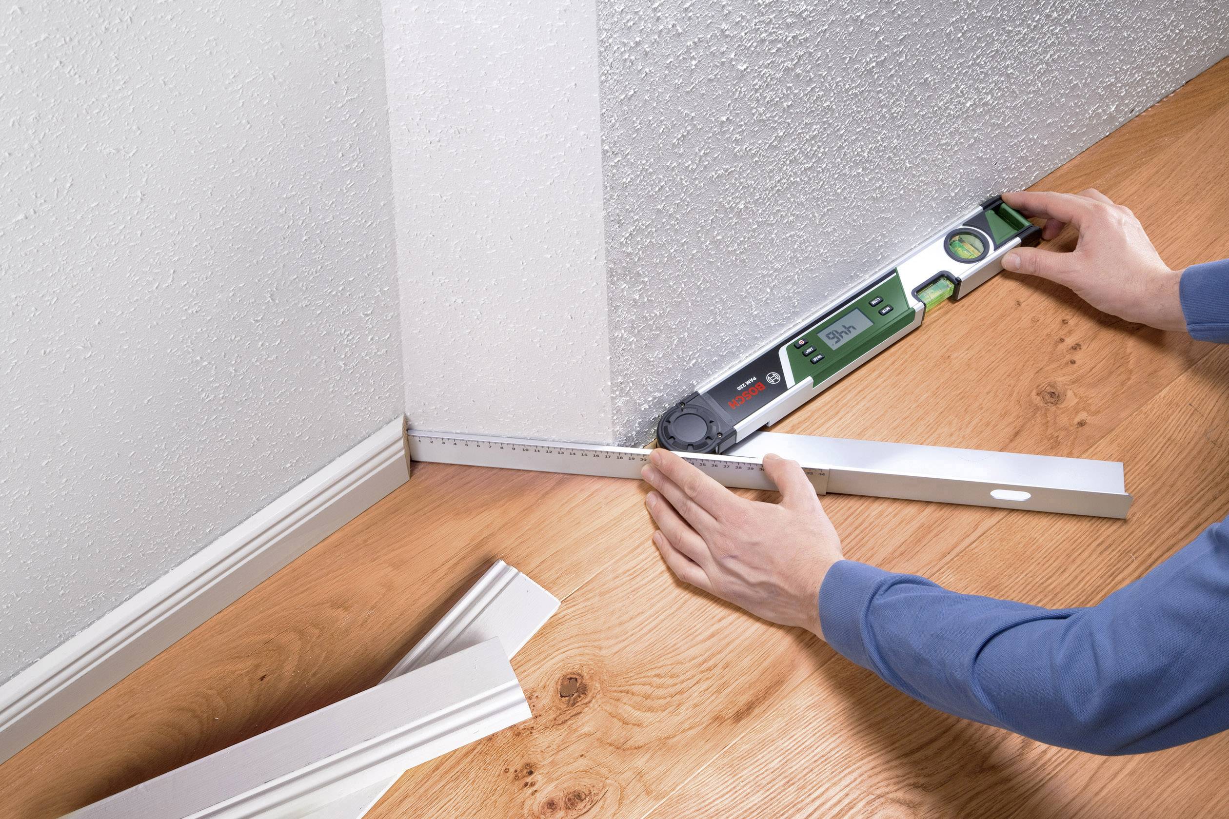 Eine Person misst mit einem digitalen Winkelmesser den rechten Winkel zwischen Wand und Boden auf einem hölzernen Fußboden.