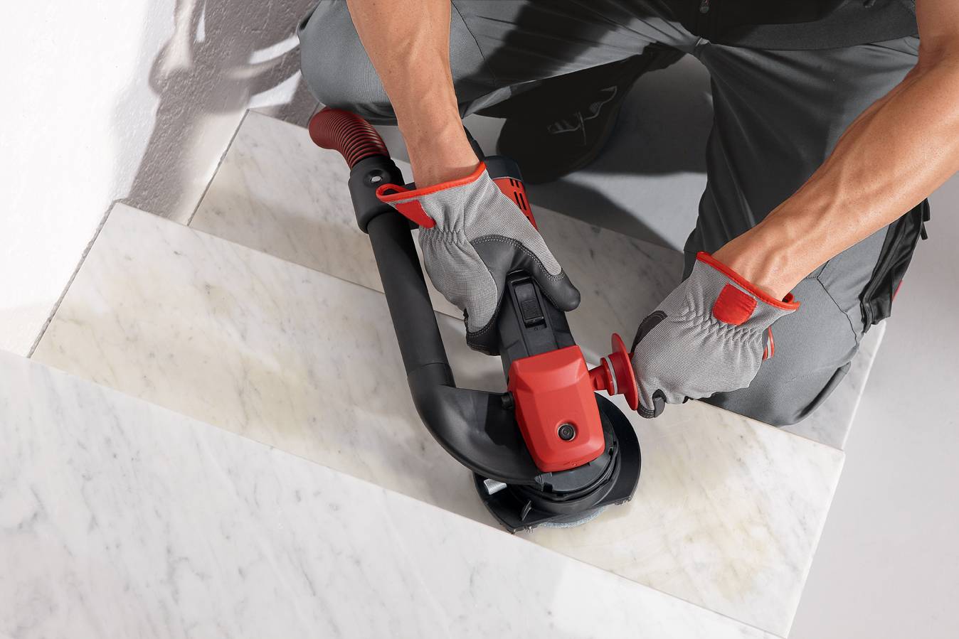Eine Person schneidet mit einem Winkelschleifer etwas auf einer Marmortreppe. Trägt graue Handschuhe und Arbeitsbekleidung.