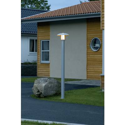 Eine leuchtende Straßenlaterne steht auf einem Rasenstück vor einem modernen Holzhaus mit runden und quadratischen Fenstern.