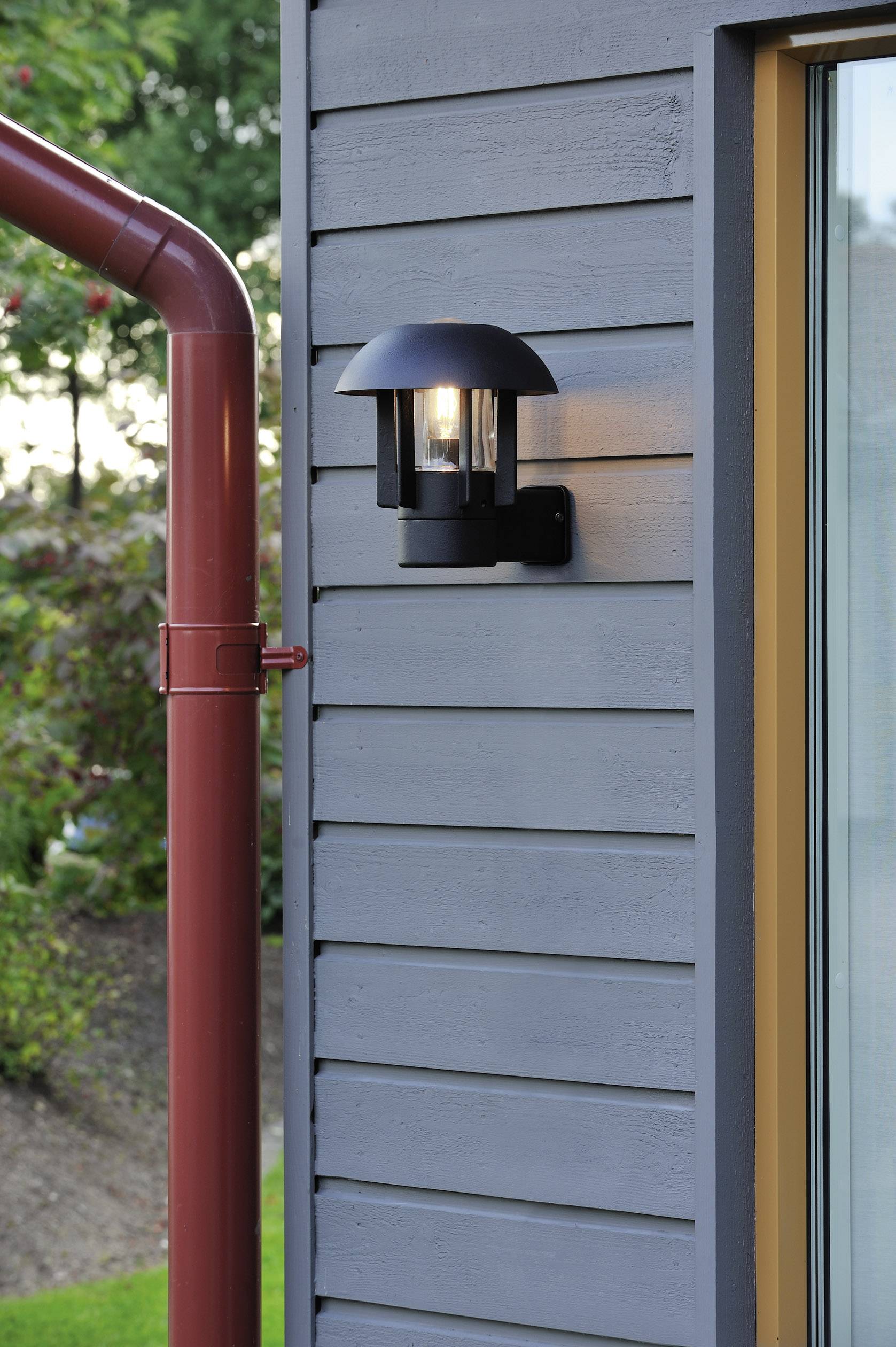 Außenlampe leuchtet an einer modernen, grau gestrichenen Hauswand neben einem roten Fallrohr. Im Hintergrund ist Grün zu sehen.