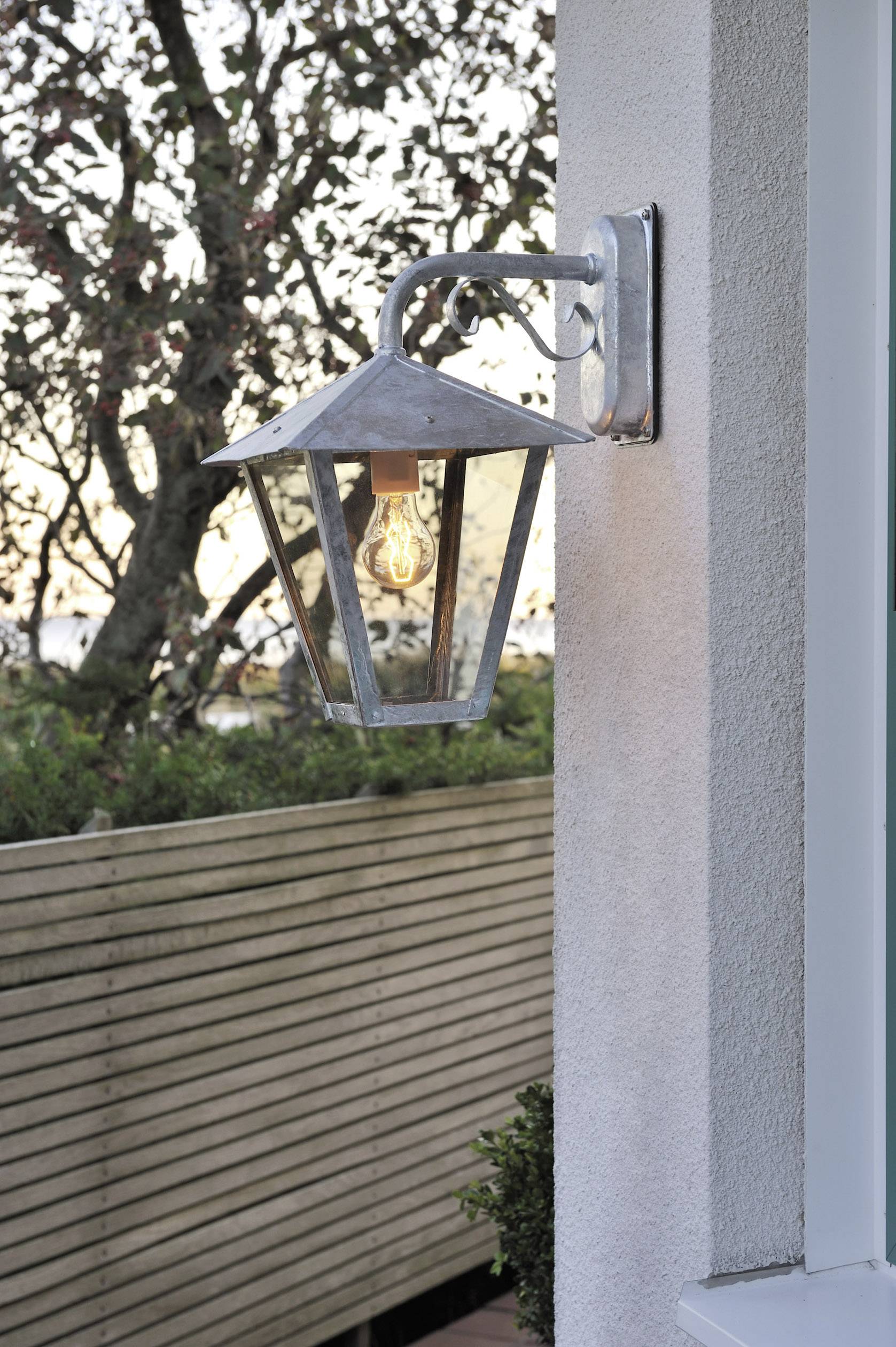 Wandleuchte aus Metall mit Vintage-Glühbirne an der Hauswand, im Hintergrund ein Holzzaun und Bäume bei Sonnenuntergang.