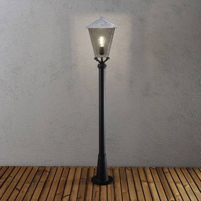 Stehlampe im antiken Stil mit quadratischem, gläsernem Lampenschirm und Metallpfosten auf hölzernem Boden vor grauer Wand.