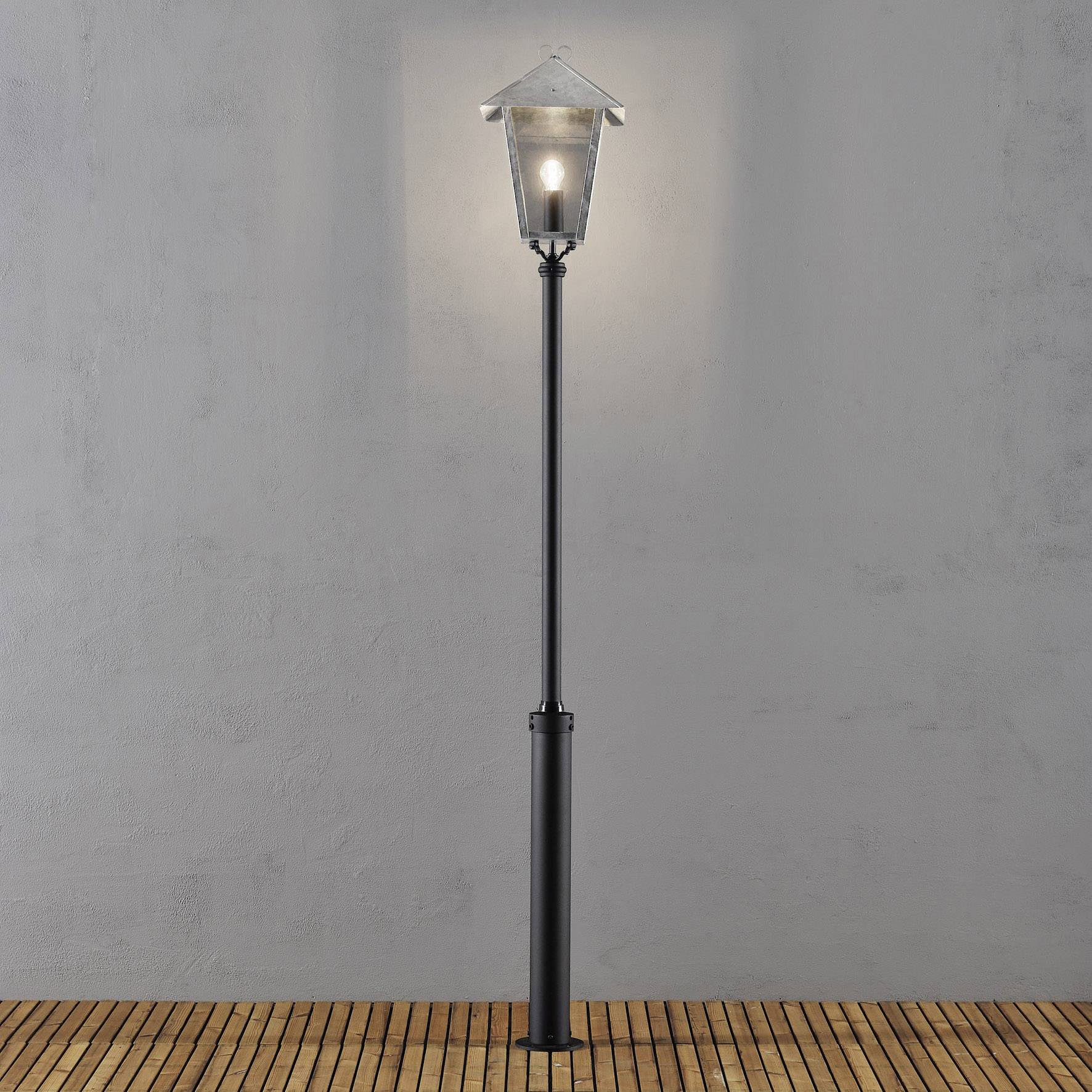 Stehlampe mit klassischem Design, leuchtet in dunkler Umgebung, steht auf einem Holzfußboden vor grauer Wand.
