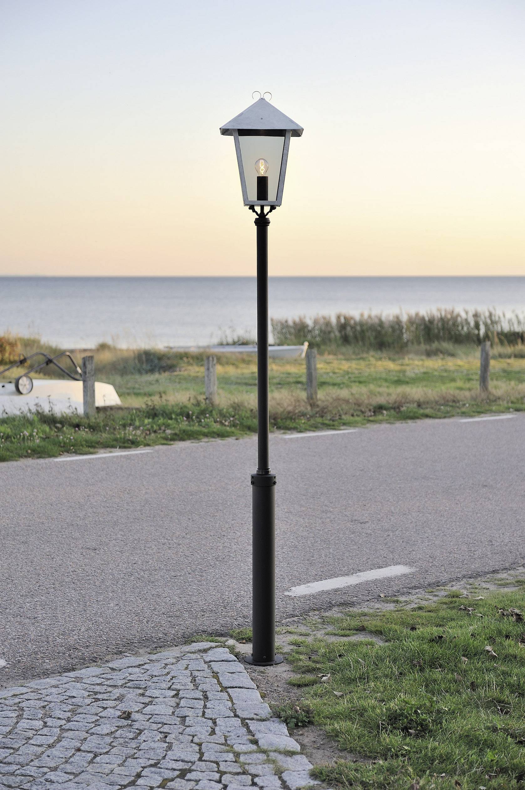 Eine Laterne steht neben einem Kiesweg am Meer. Im Hintergrund ist ein ruhiger, farbenfroher Sonnenuntergang mit wolkenlosem Himmel.