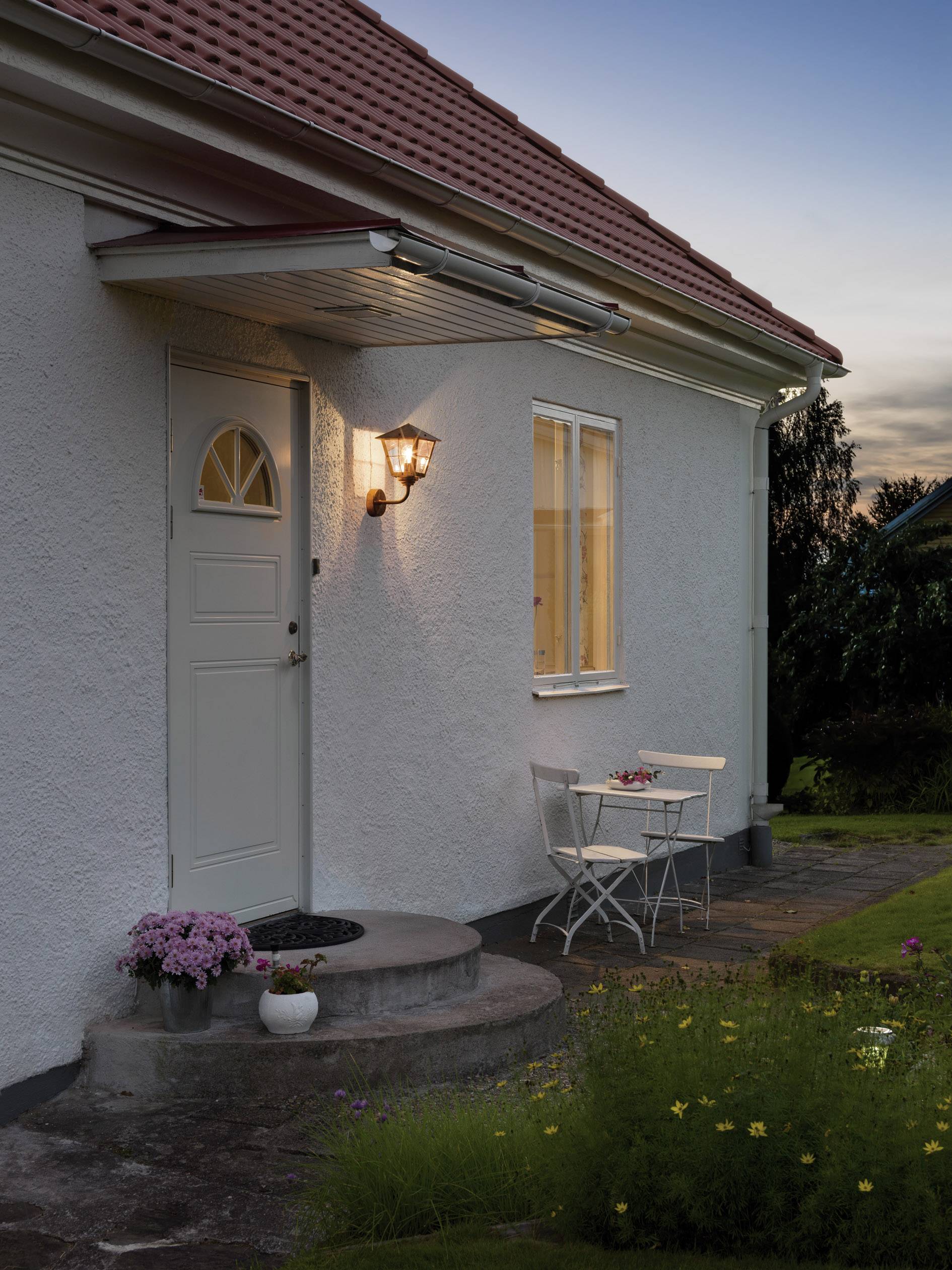 Ein weißes Haus mit einer leuchtenden Außenlampe neben der Eingangstür, einem runden Stufenabsatz und Gartenmöbeln in der Abenddämmerung.