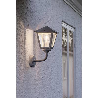 Wandleuchte an einer grauen Mauer, eingeschaltet in der Dämmerung, beleuchtet einen Bereich neben einem Fenster.
