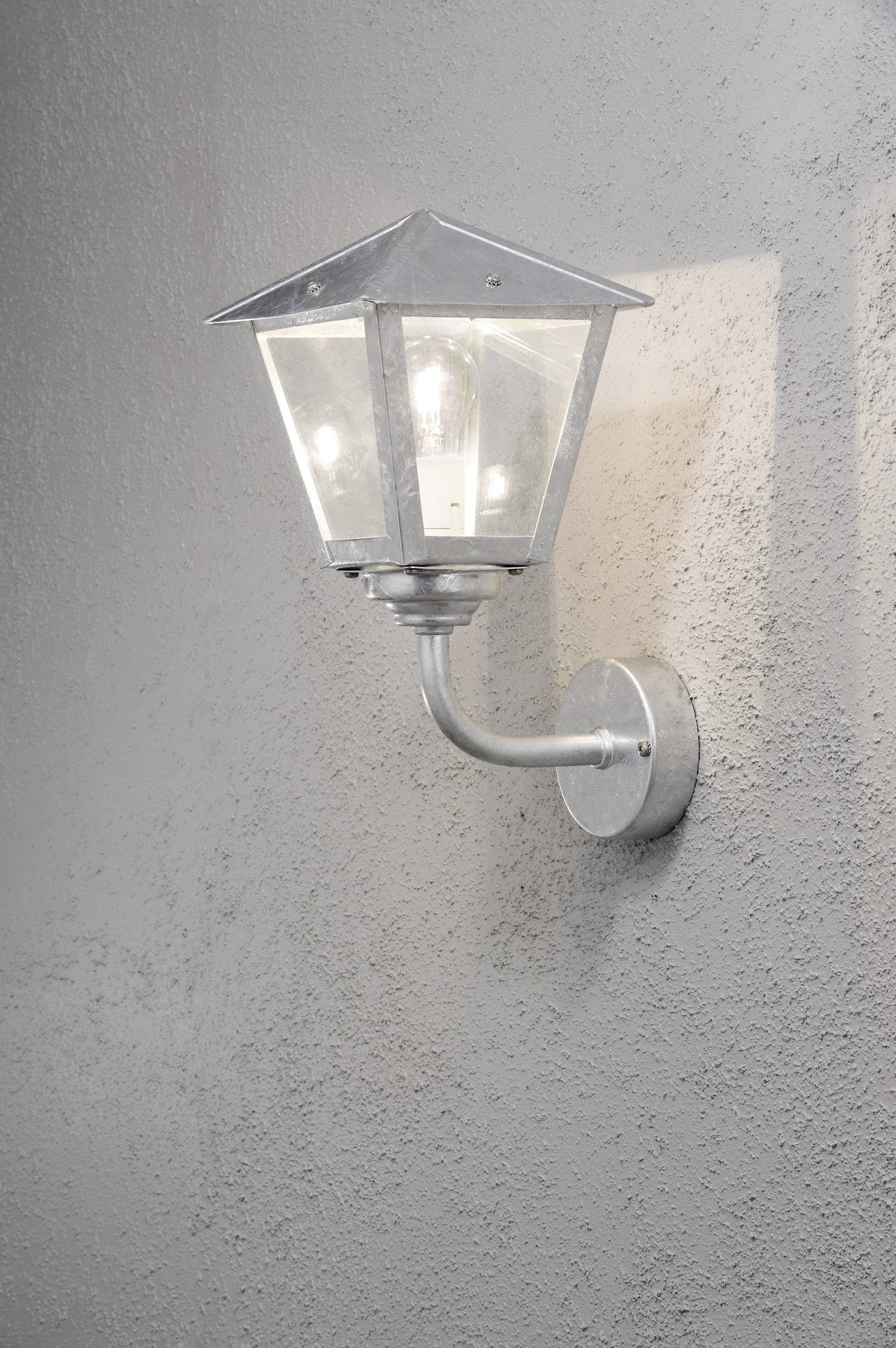Silberne Wandleuchte im Laternenstil an einer grauen Wand montiert, mit klarem Glas und sichtbarem Leuchtmittel.