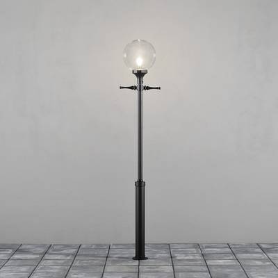 Eine schwarze Straßenlaterne mit einer kugelförmigen, transparenten Glühbirne auf einem gepflasterten Boden vor einer grauen Wand.