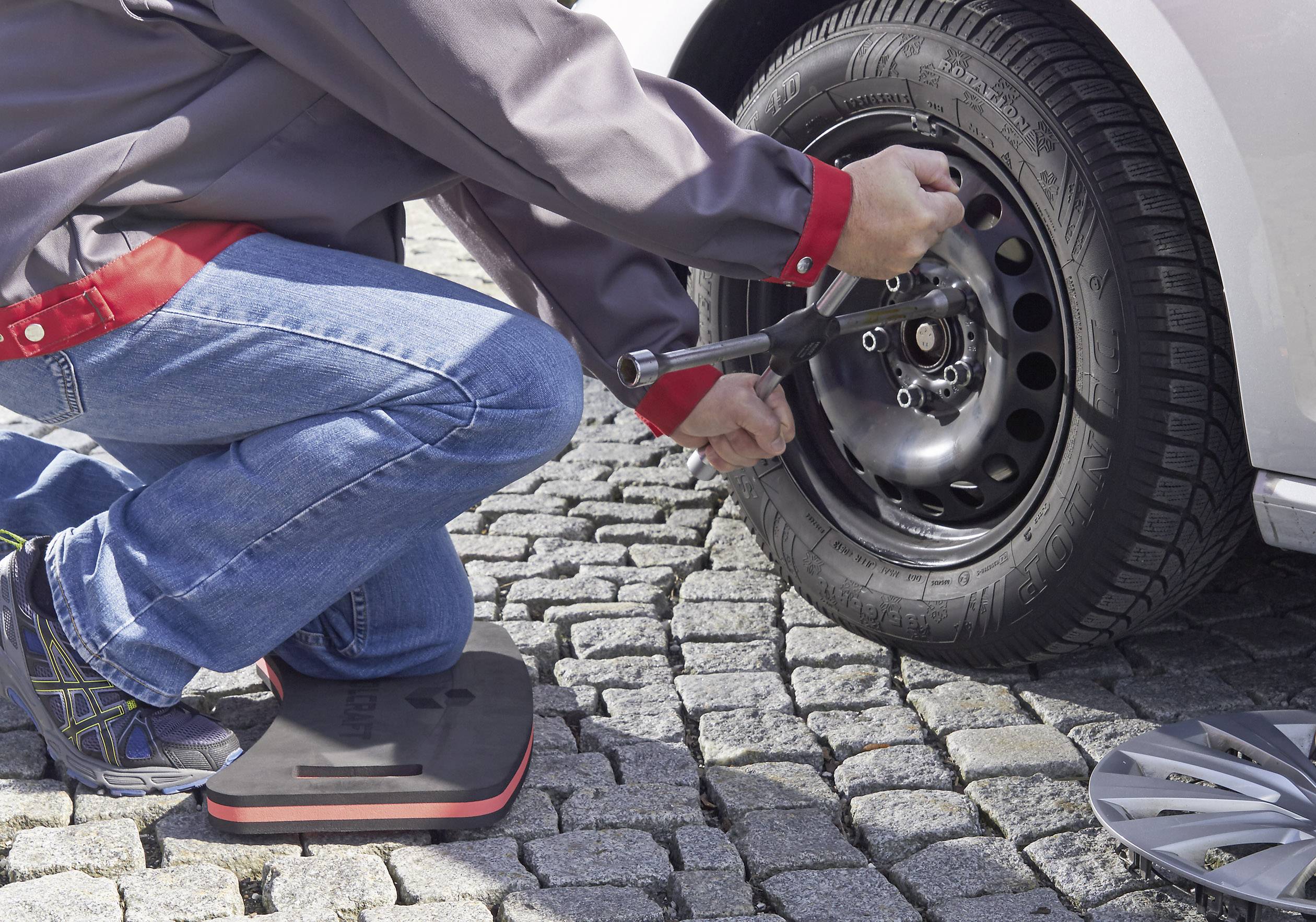 Eine Person wechselt ein Autoreifen auf einem gepflasterten Weg, verwendet dazu einen Schraubenschlüssel und trägt Freizeitkleidung.
