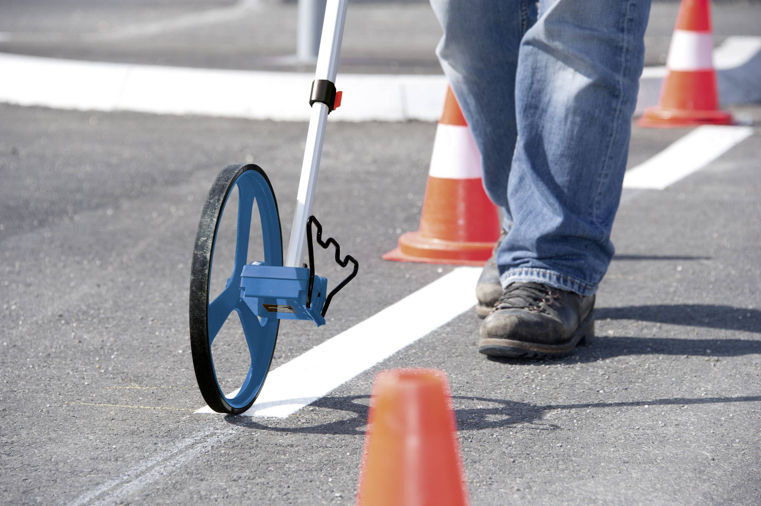 Eine Person misst eine weiße Linie auf einer Straße mit einem Messrad, umgeben von Verkehrskegeln.