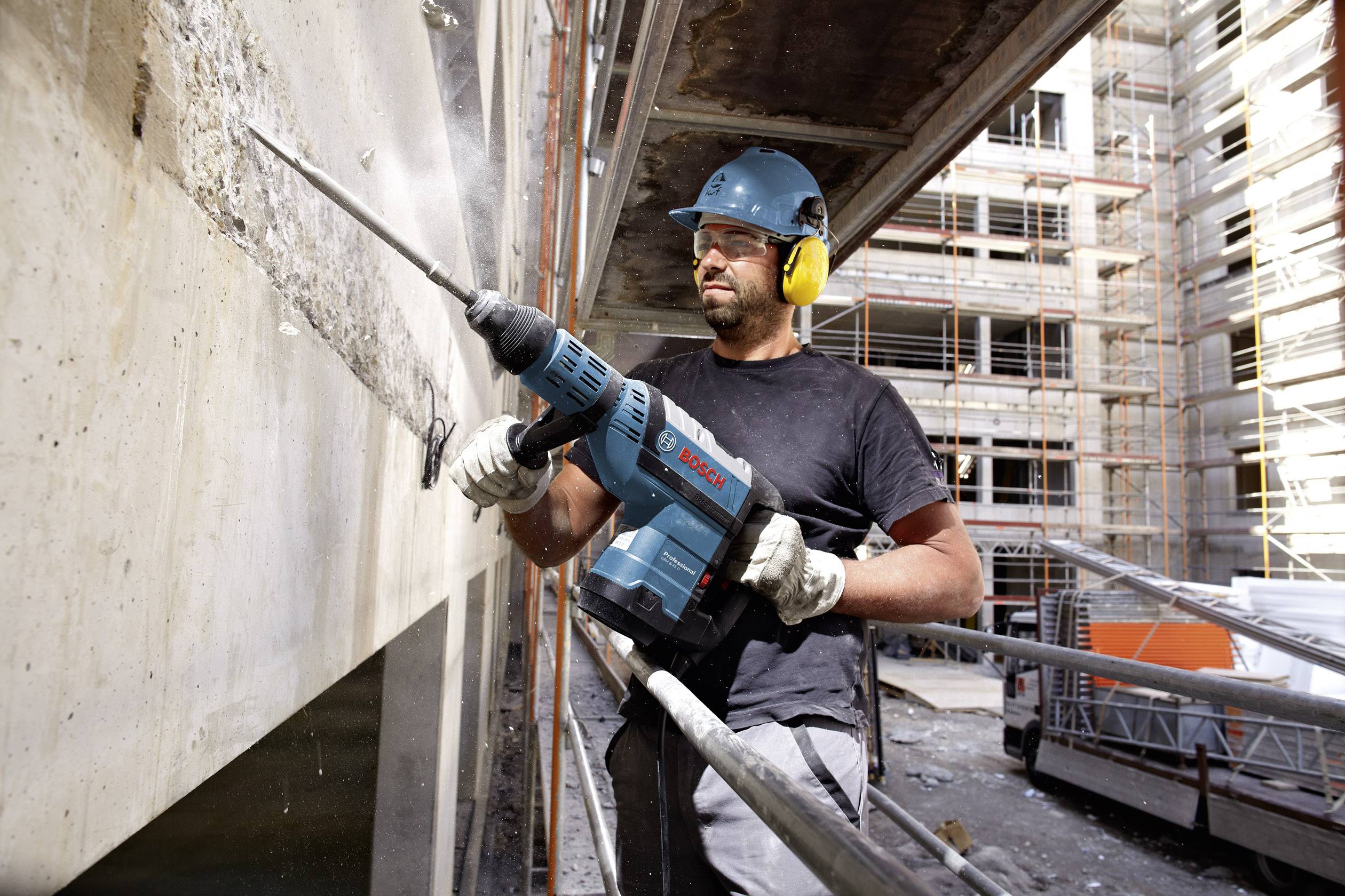 Ein Bauarbeiter trägt Schutzkleidung und nutzt einen Bohrhammer, um eine Betonwand auf einer Baustelle zu bearbeiten. Im Hintergrund sind Baugerüste sichtbar.