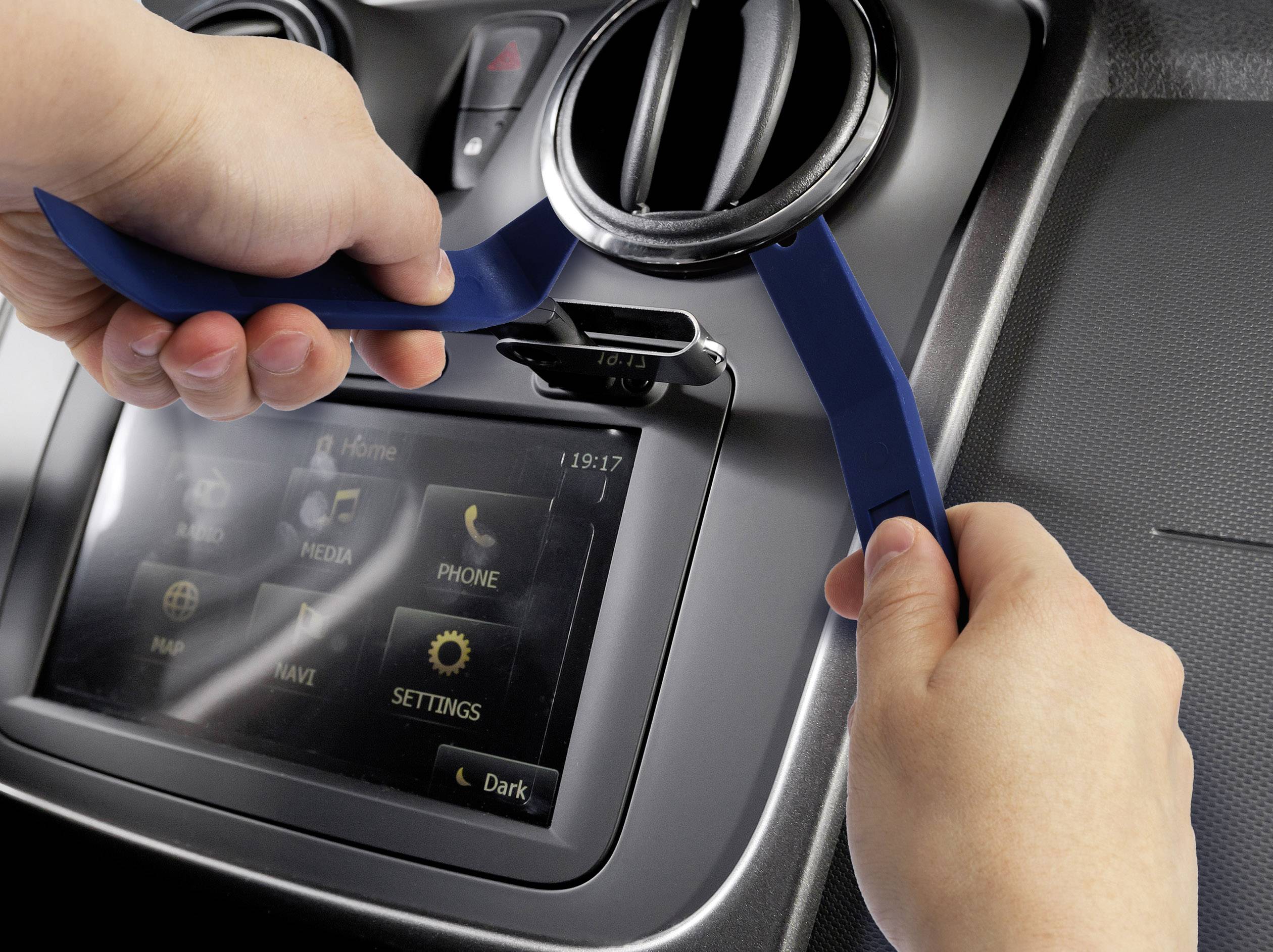 'Zwei Hände verwenden blaue Plastikwerkzeuge, um ein Lüftungsgitter im Auto zu entfernen. Dahinter befindet sich ein Bildschirm.'