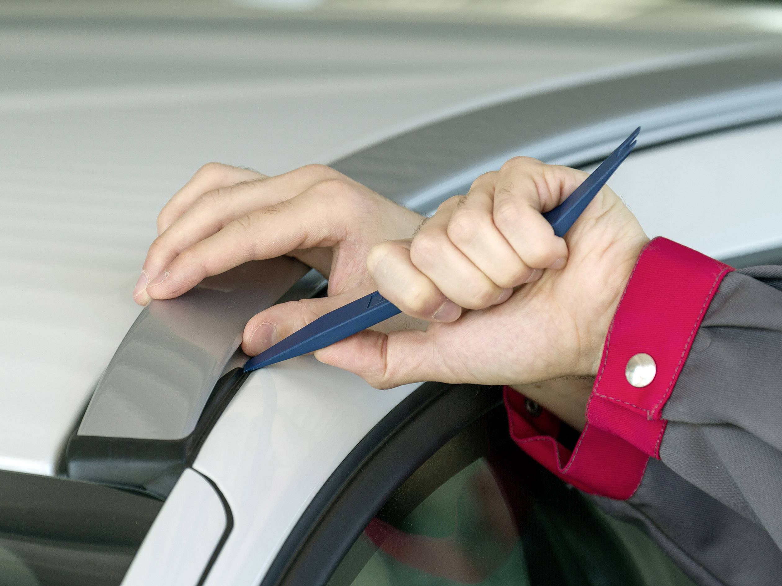 Eine Person verwendet ein blaues Plastikwerkzeug, um die Dichtungen eines Autofensters sorgfältig zu öffnen.