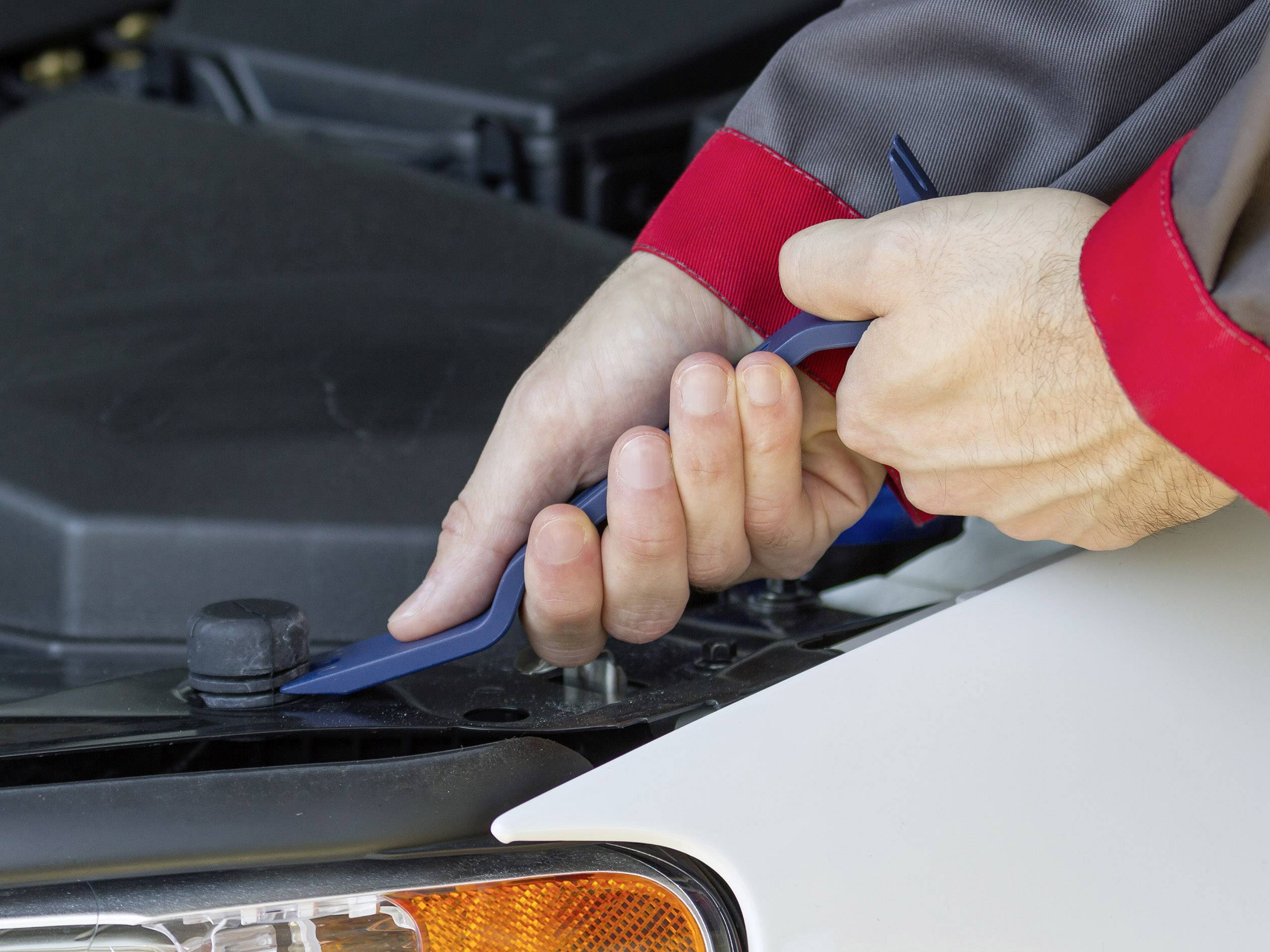 Hände eines Mechanikers benutzen ein Kunststoffwerkzeug, um die Motorhaube eines Autos zu öffnen. Das Auto ist weiß.