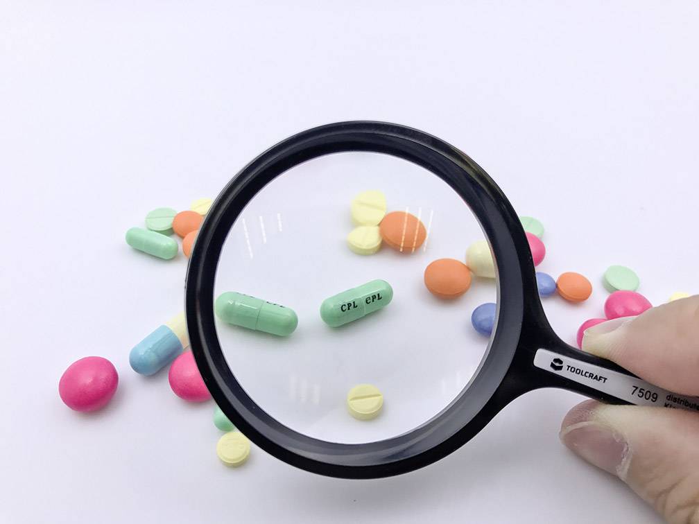 Vergrößerungsglas über bunte Tabletten, darunter grüne mit Aufschrift 'CPL'. Darum verstreut weitere Tabletten in verschiedenen Farben.