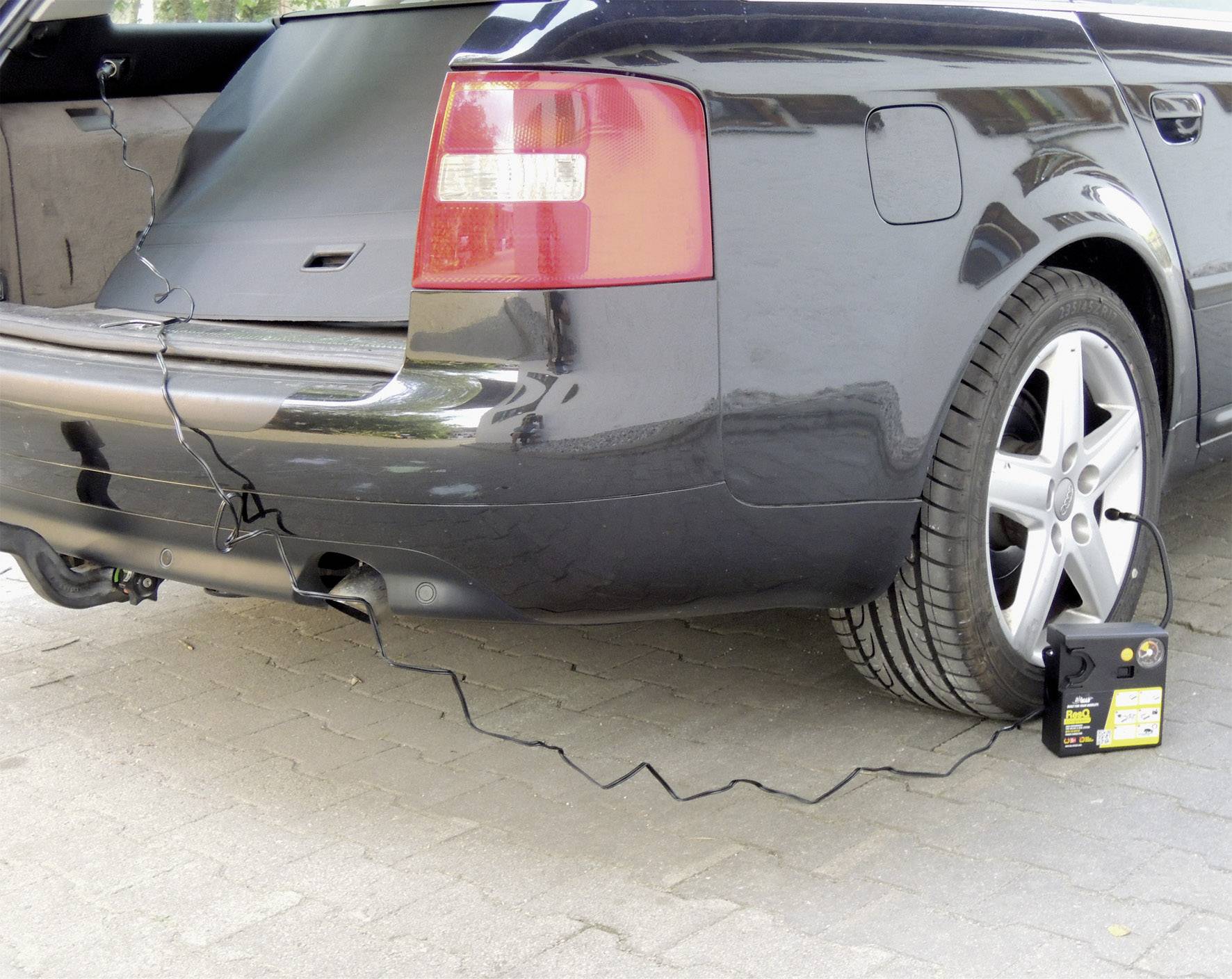 Schwarzes Auto mit geöffnetem Kofferraum. Ein Ladegerät ist an die Autobatterie angeschlossen und liegt neben dem Hinterrad.