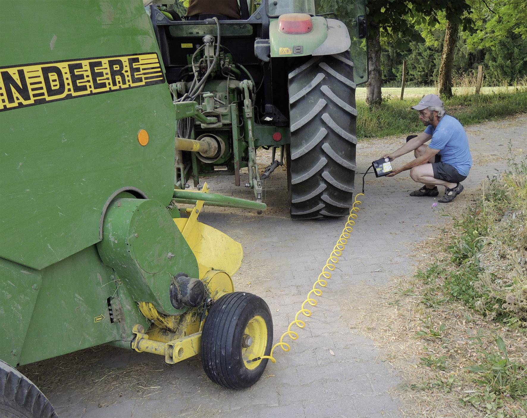 Ein Mann pumpt einen Reifen einer grünen landwirtschaftlichen Maschine auf. Die Maschine steht auf einem gepflasterten Weg.