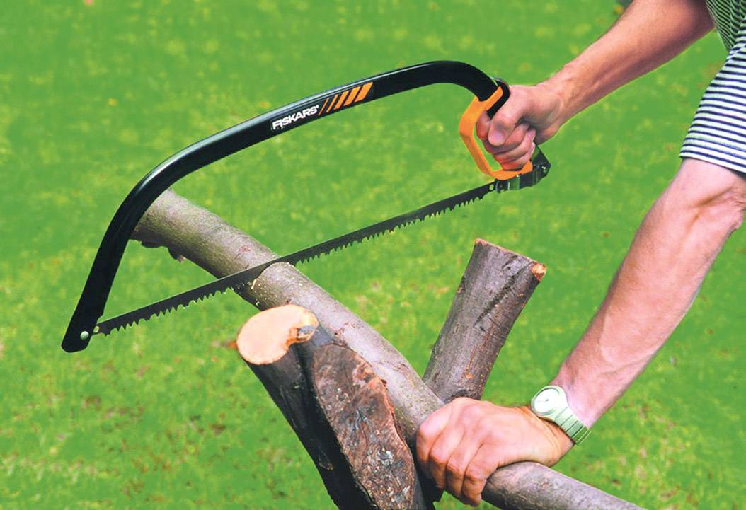 Eine Person sägt mit einer Bügelsäge einen Ast auf einem Rasen. Sie hält den Ast mit der linken Hand fest.