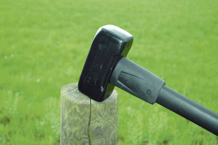 Ein schwarzer Hammerkopf auf einem Holzpfosten in einer grünen Wiese. Der Hammer zeigt Risse im Holz, deutet auf Pfostenarbeiten hin.