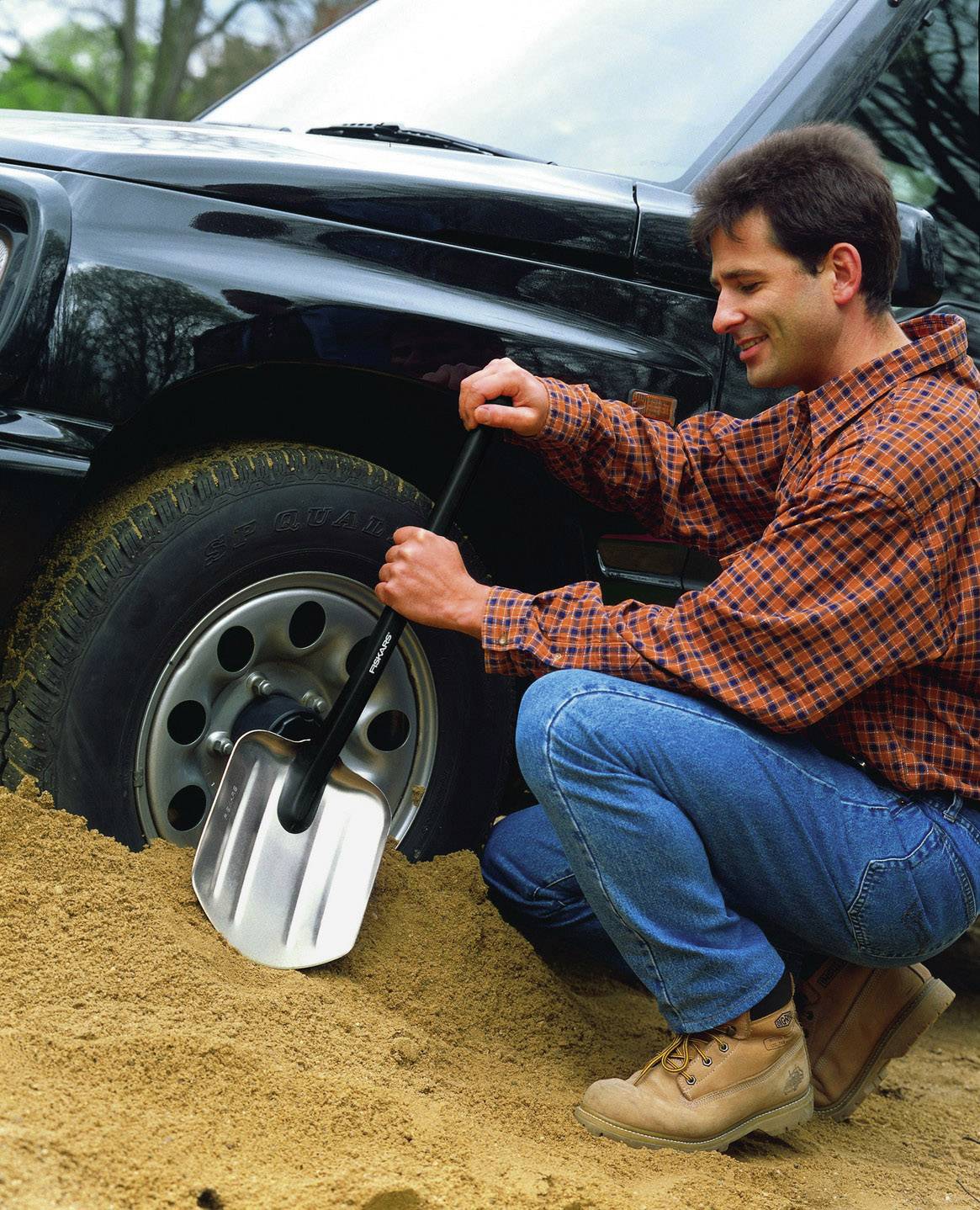 Ein Mann in Arbeitskleidung gräbt mit einer Schaufel Sand neben einem Autoreifen aus. Das Auto steht in sandigem Gelände.