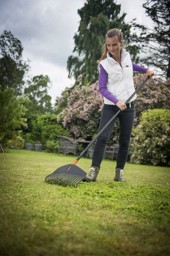 Eine Person harkt im Garten, umgeben von Bäumen und blühenden Sträuchern, unter einem bewölkten Himmel.