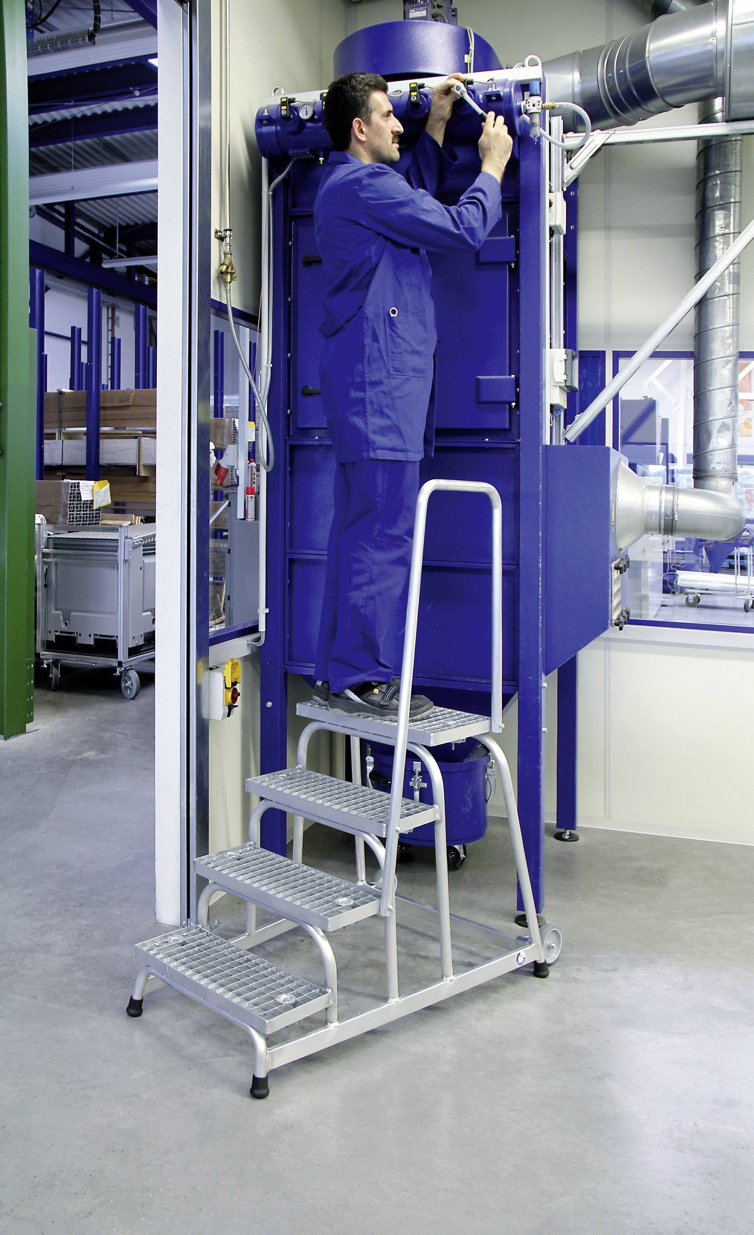 Ein Techniker im blauen Overall steht auf einer Leiter und arbeitet an einer großen blauen Maschine in einer industriellen Umgebung.
