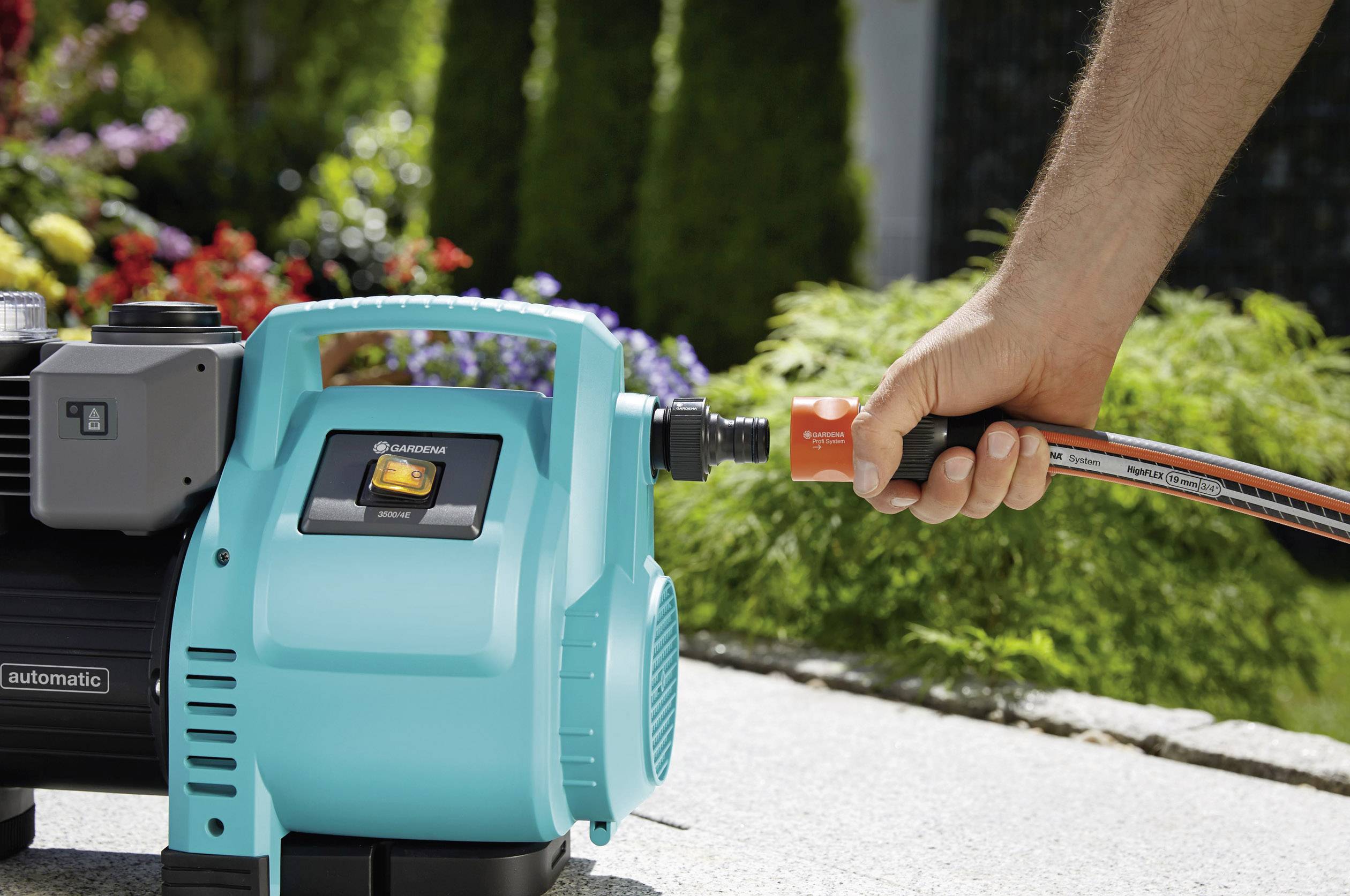 Eine Person verbindet einen Gartenschlauch mit einer blauen Wasserpumpe in einem Garten. Im Hintergrund sind Pflanzen sichtbar.