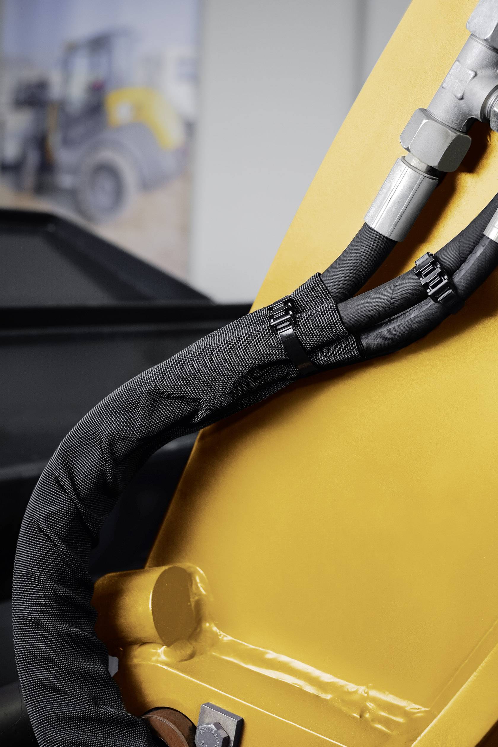 Nahaufnahme eines gelben Baumaschinenarms mit schwarzen Hydraulikschläuchen, die durch Metallklemmen gesichert sind. Im Hintergrund unscharf.