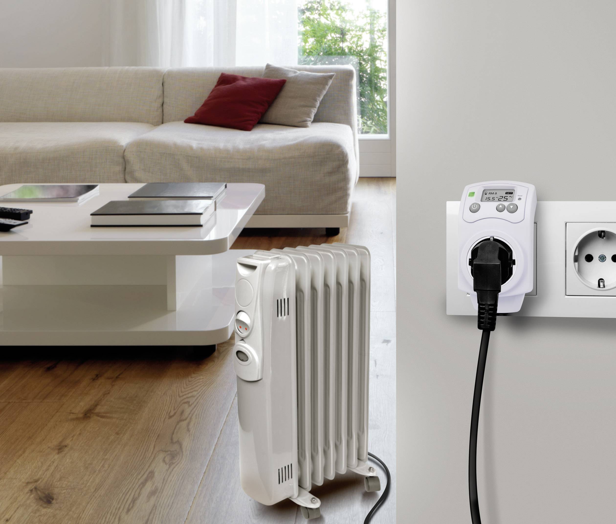 Ein elektrischer Heizkörper steht in einem Wohnzimmer neben einem Steckdosen-Timer an der Wand. Im Hintergrund ein Sofa mit Kissen.
