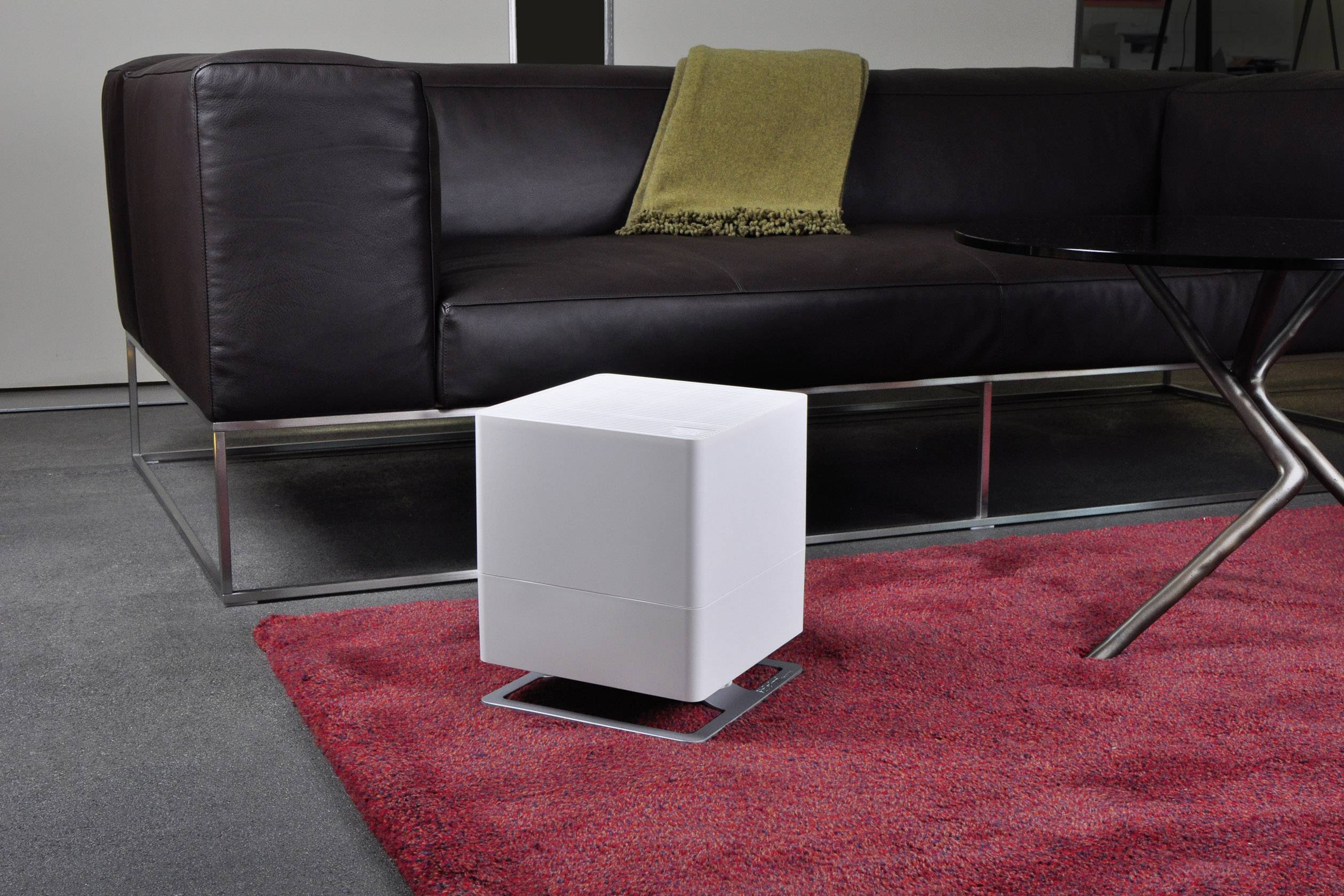 Ein weißer, würfelförmiger Lautsprecher steht auf einem roten Teppich vor einem braunen Ledersofa mit beige Decke im modernen Wohnzimmer.