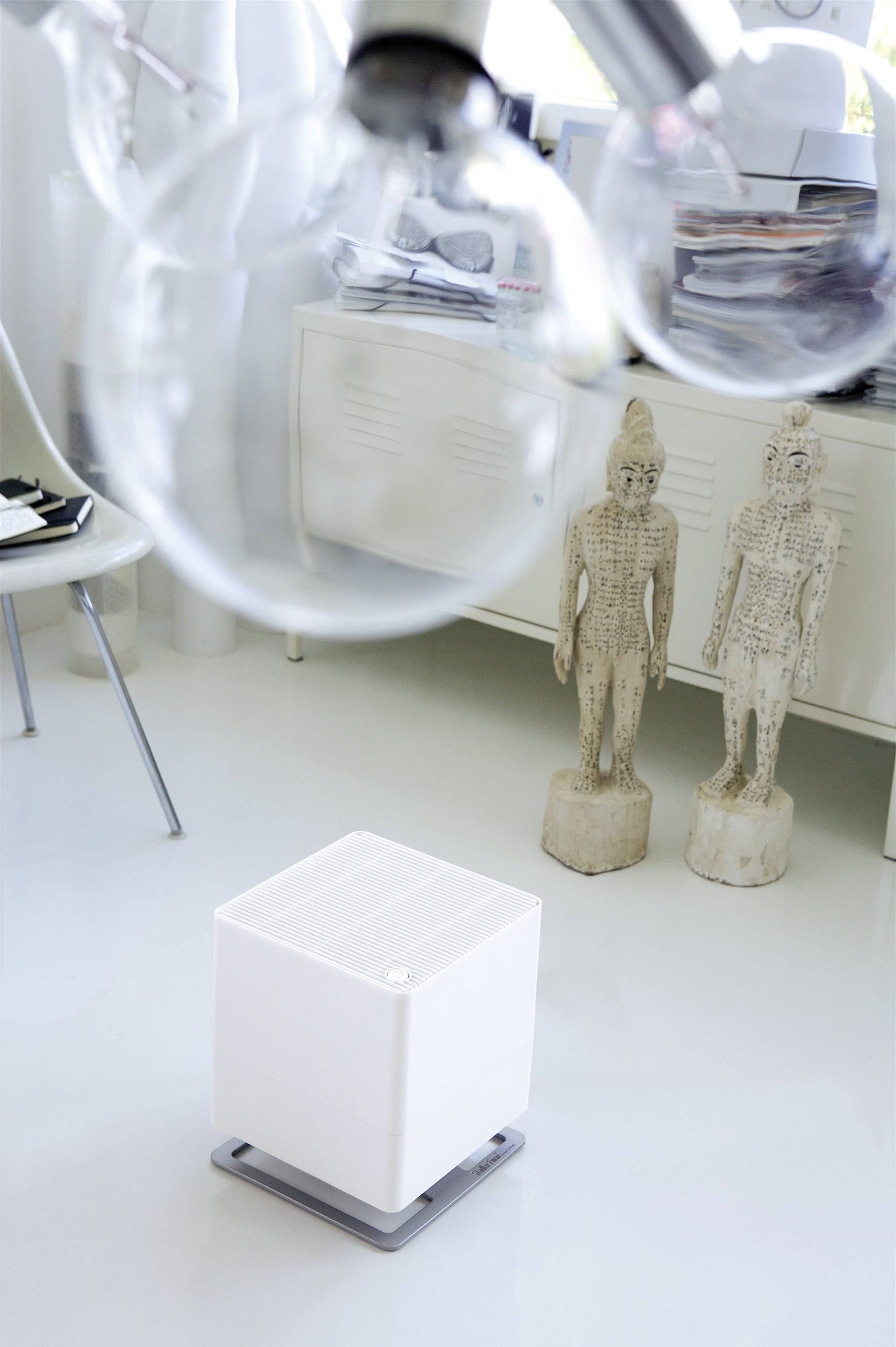 Ein weißer Luftreiniger steht auf einem hellen Boden in einem modernen Raum. Im Hintergrund sind zwei Statuen zu sehen.
