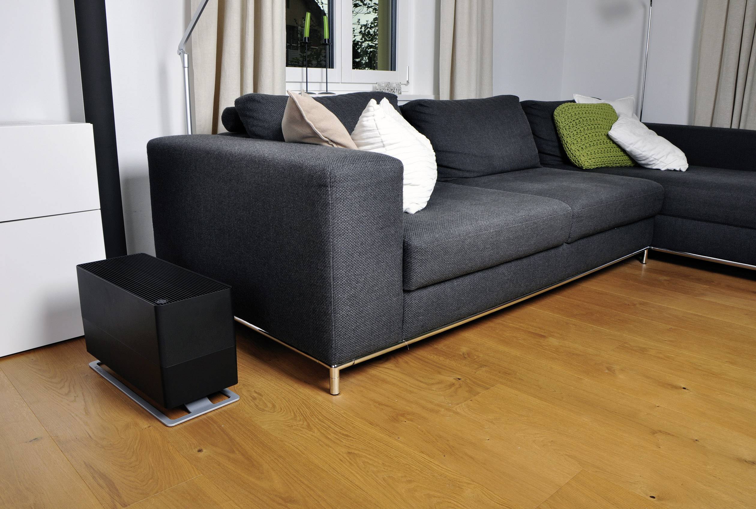 Dunkelgraues Sofa in einem modern eingerichteten Wohnzimmer mit Holzfußboden. Ein schwarzes Gerät steht links neben dem Sofa.