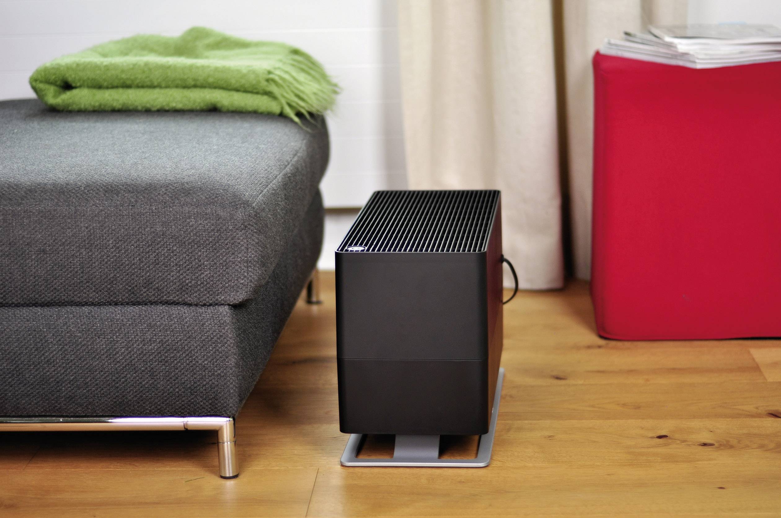 Ein moderner Luftbefeuchter steht auf einem Holzboden neben einem grauen Sofa mit grünem Tuch und einem roten Hocker mit Zeitungen.